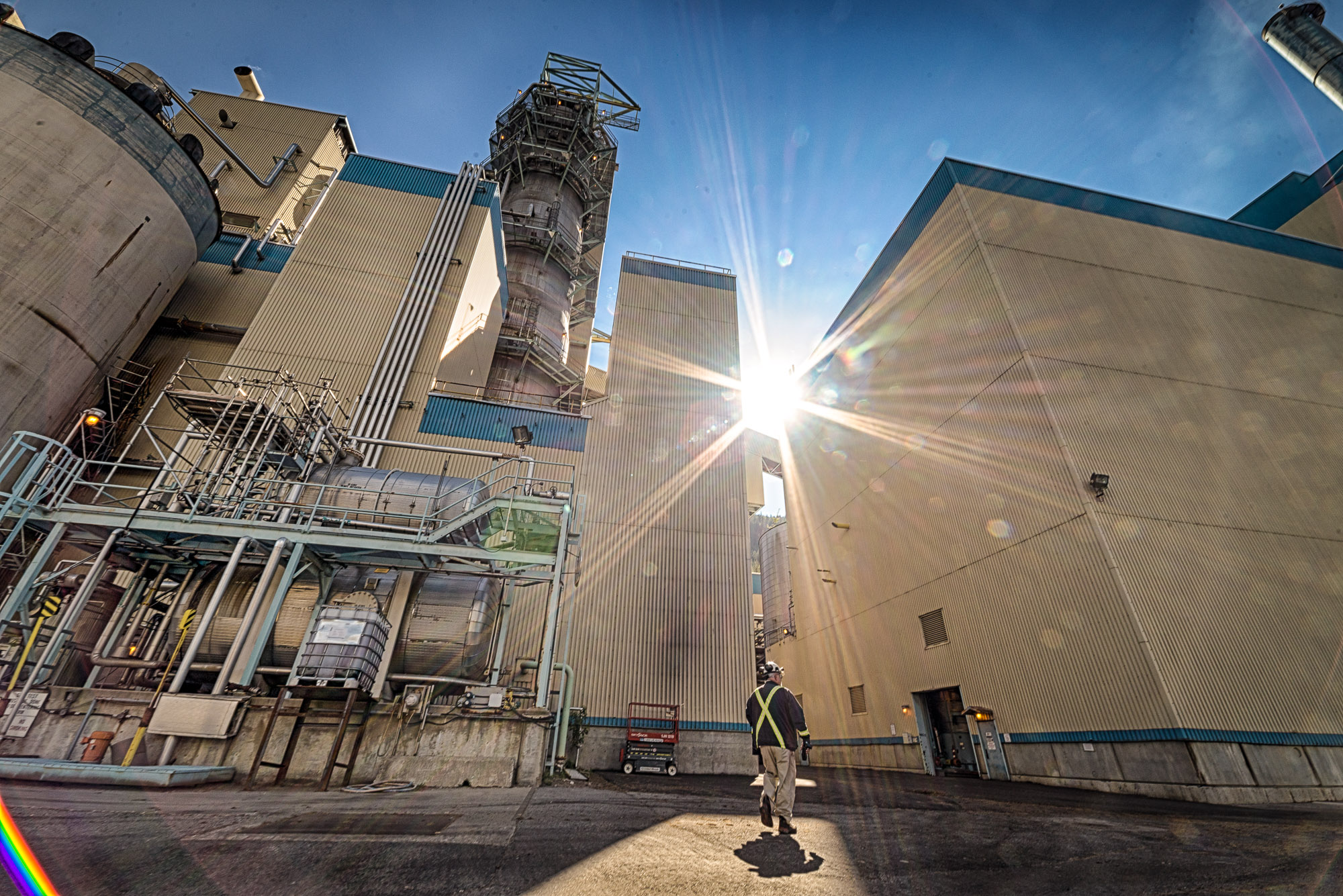 Worker in high-vis walks between towering plant structures as sun flares between buildings at a Kootenays industrial site.