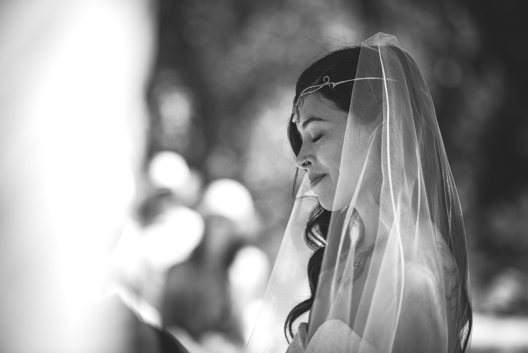 Bride closes her eyes beneath a sheer veil, soft dappled light filtering through trees behind her.